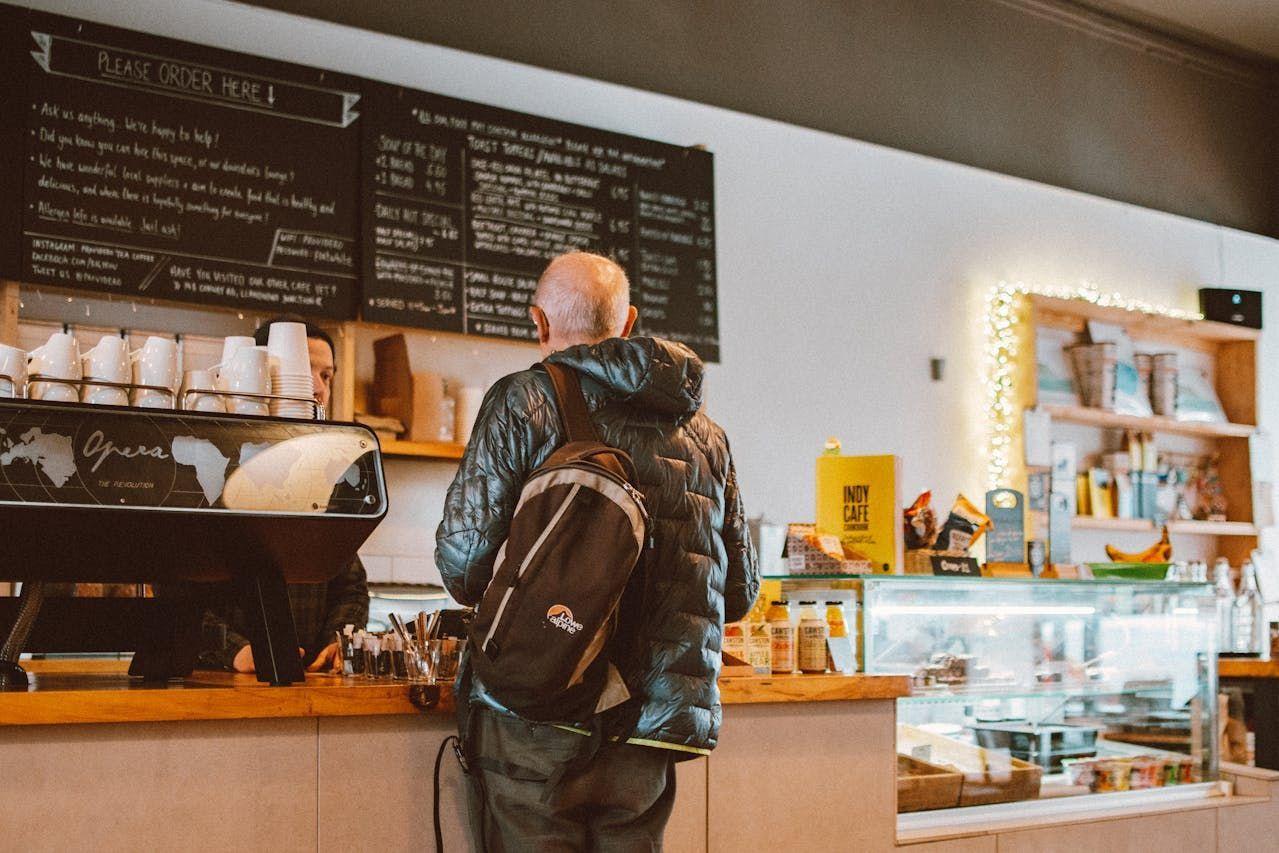 A man with a backpack stands at a coffee shop counter, looking at the menu board.