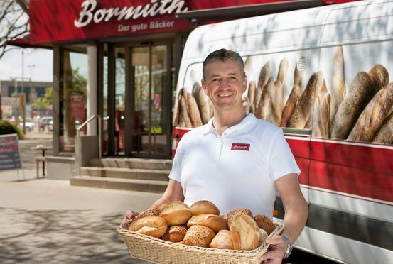 Smiling baker holding a basket of fresh bread rolls outside a Bormuth bakery.