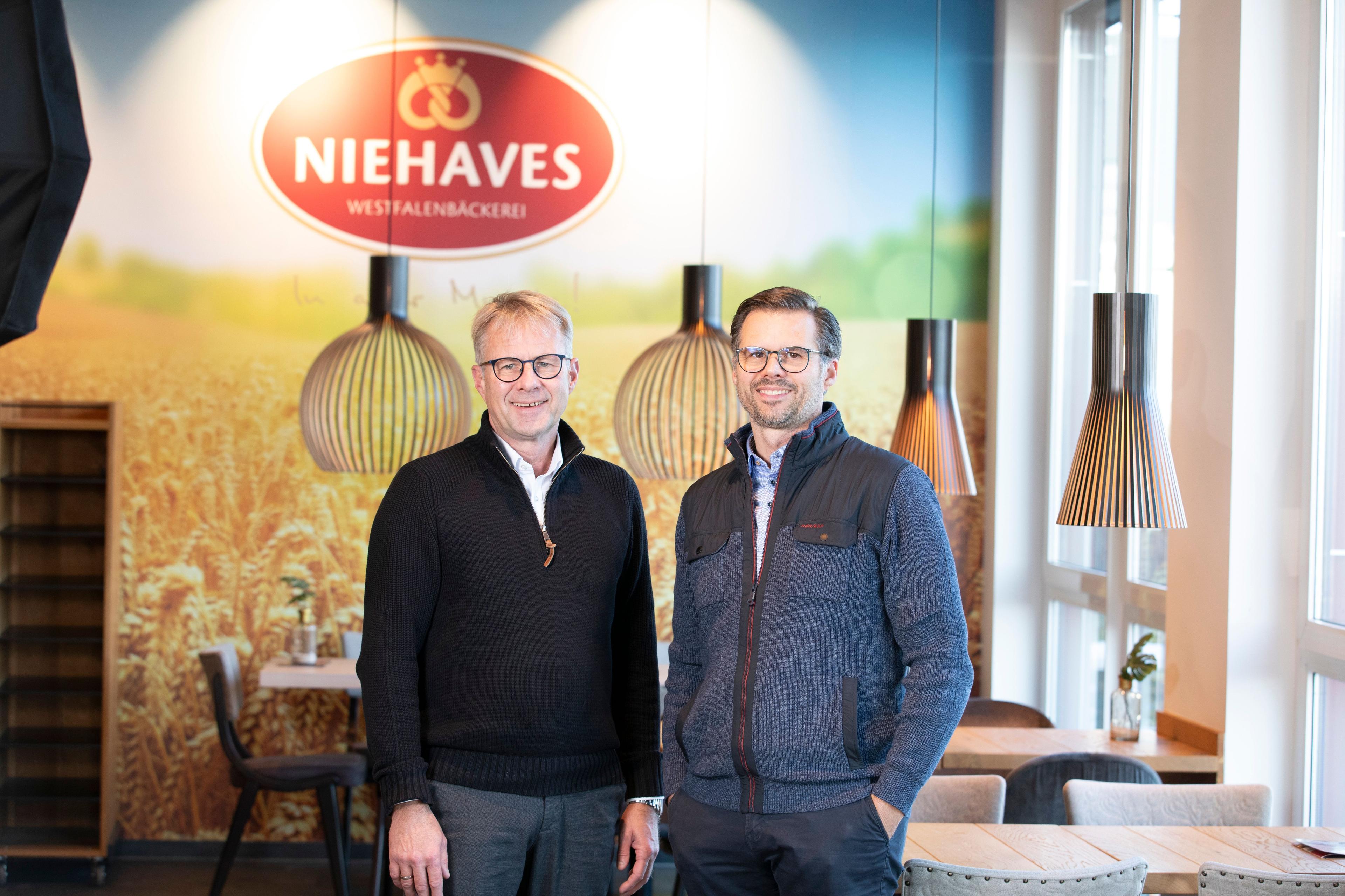 Two men stand in a modern bakery with the Niehaves brand logo on the wall.