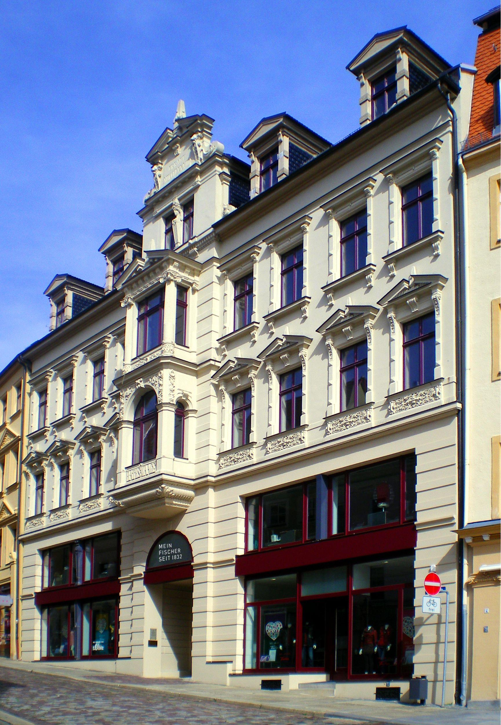 Ornate, multi-story building with light facade and ground-floor shop windows under a blue sky.