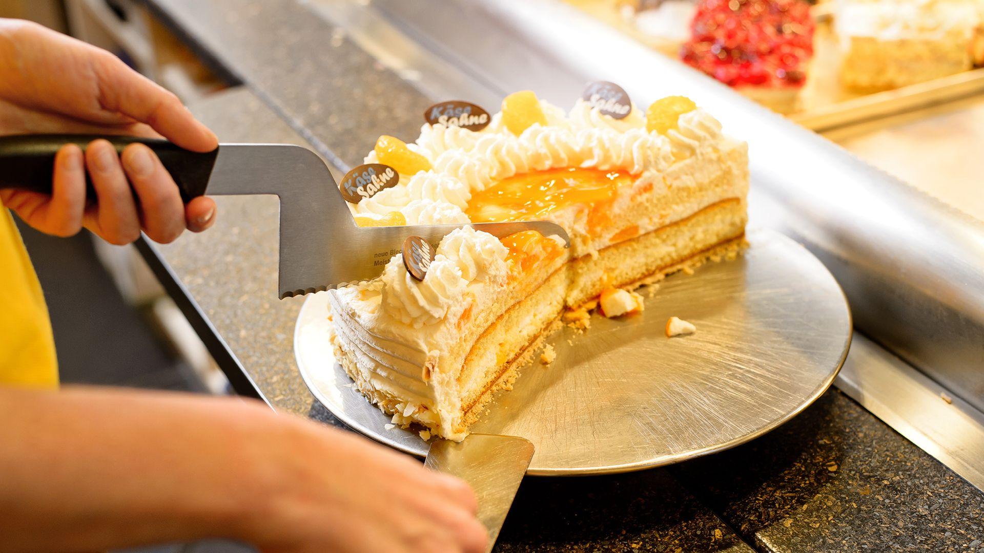 Hands cutting a slice of cream-frosted cake with fruit and chocolate garnish.