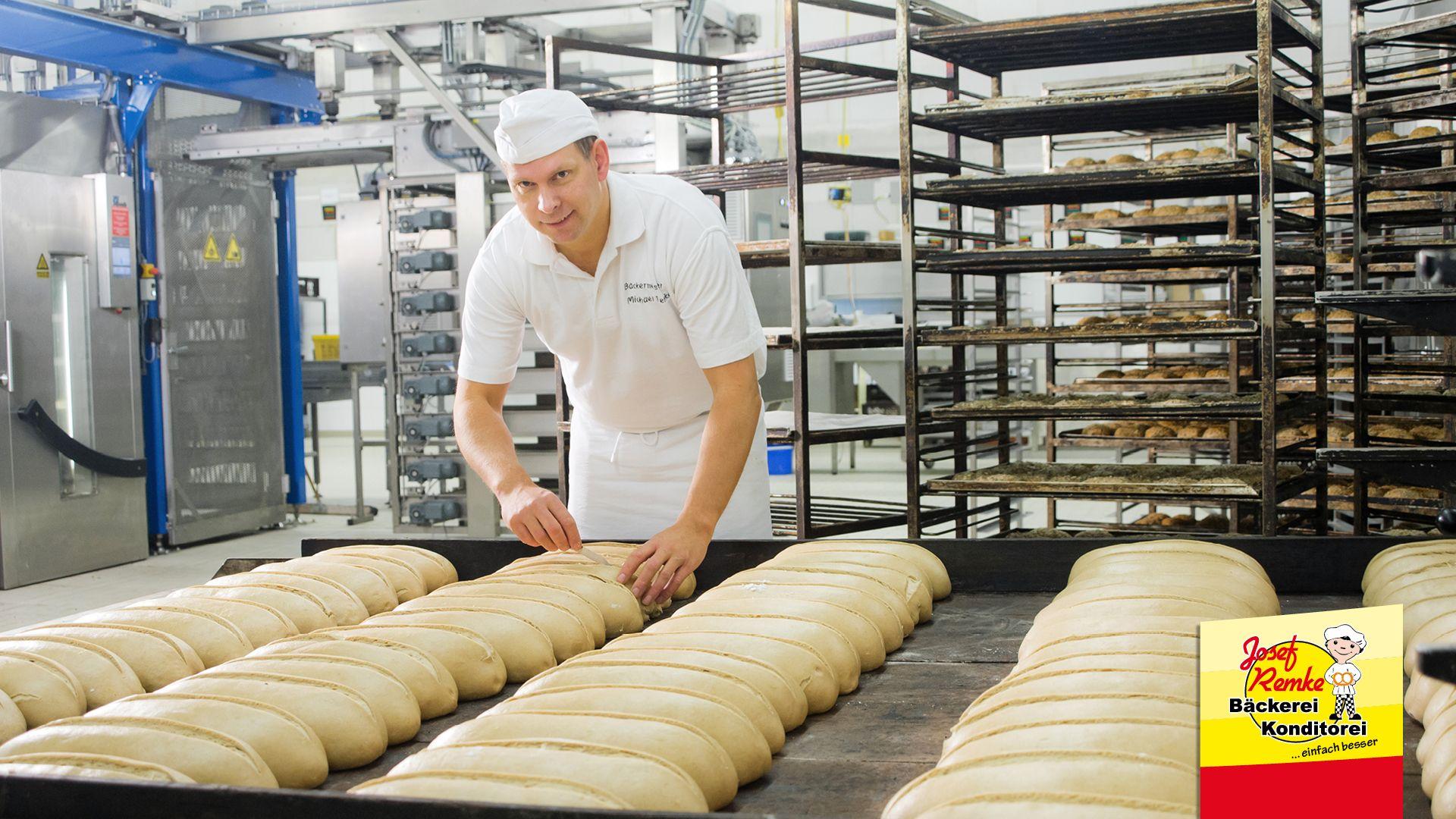 Baker arranges raw bread dough loaves on trays in a large commercial bakery.