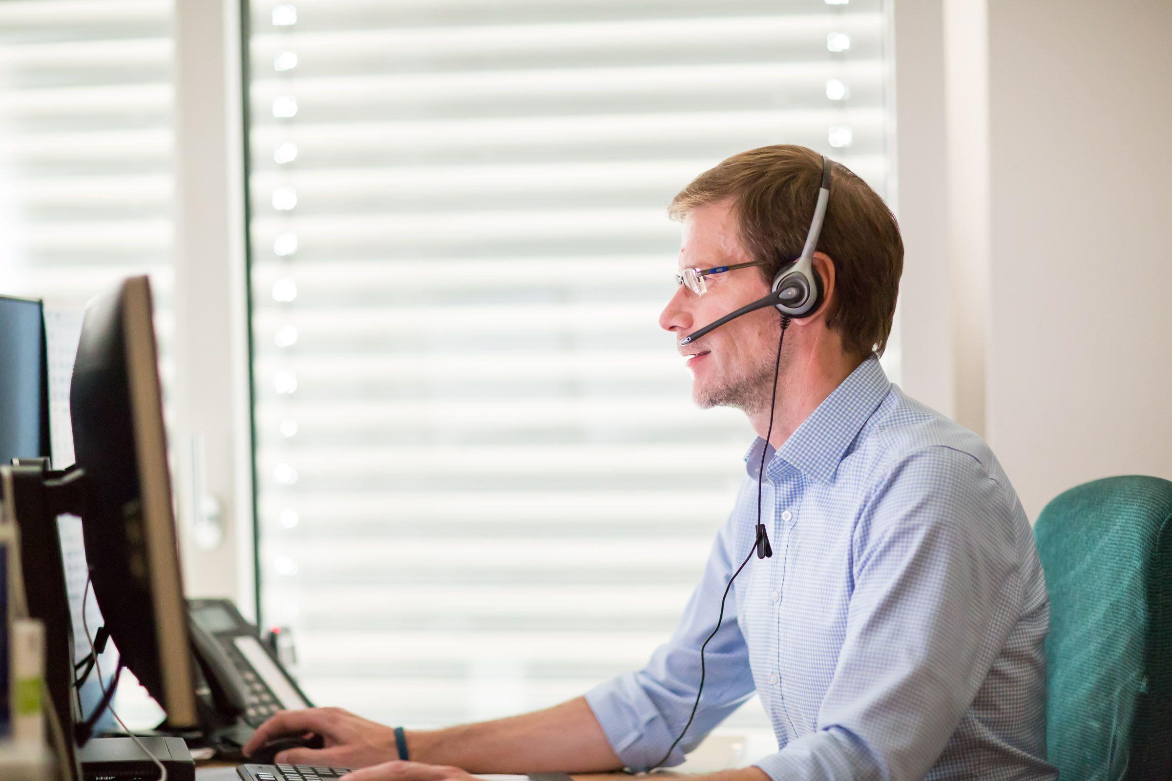 Man in headset and glasses smiling while working on computer at his desk.