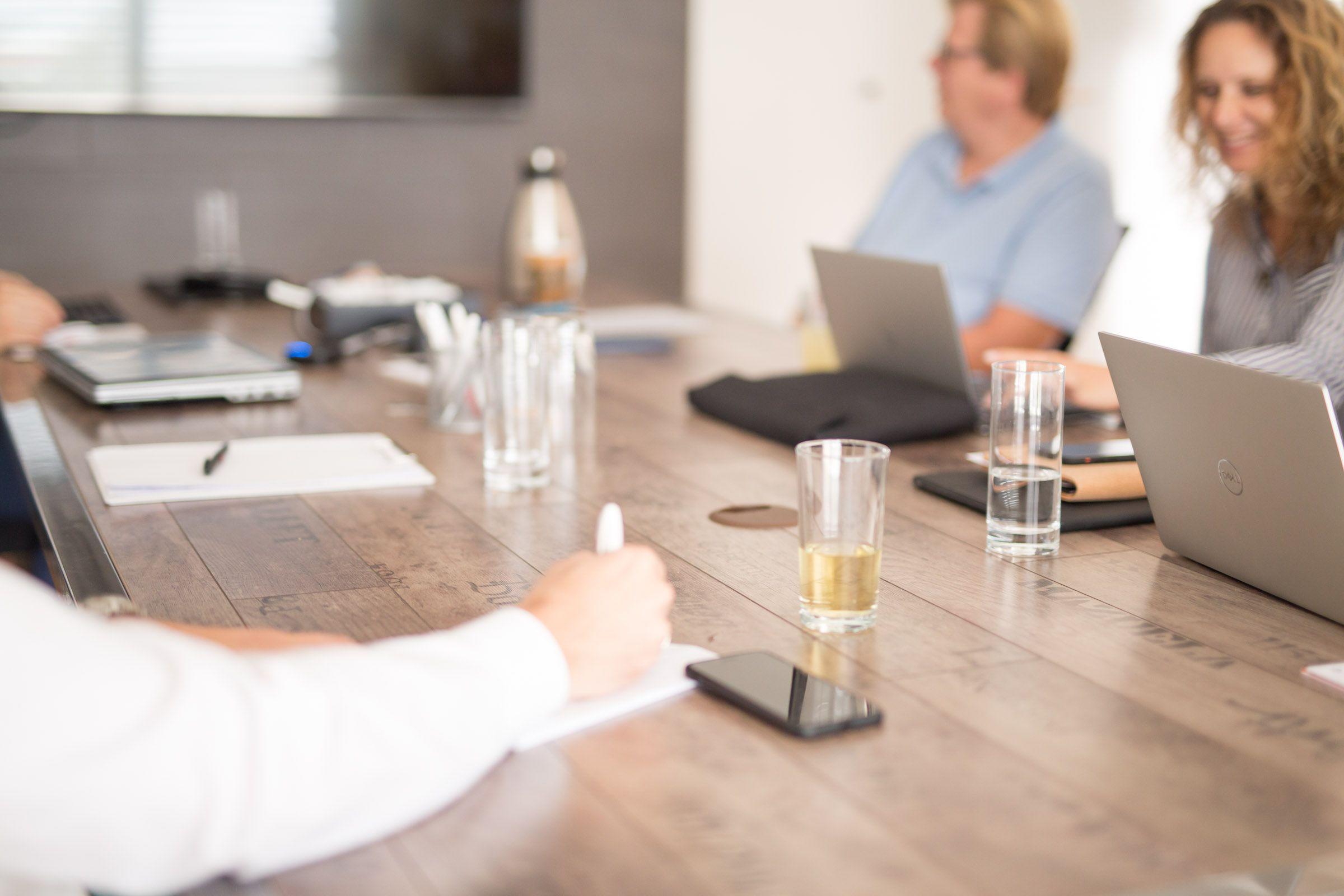 People collaborate around a wooden conference table with laptops, notebooks, and drinks.