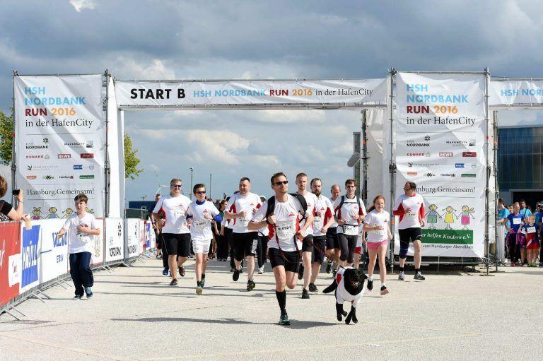 Runners and a dog start a race under a "START B" banner on a sunny day.