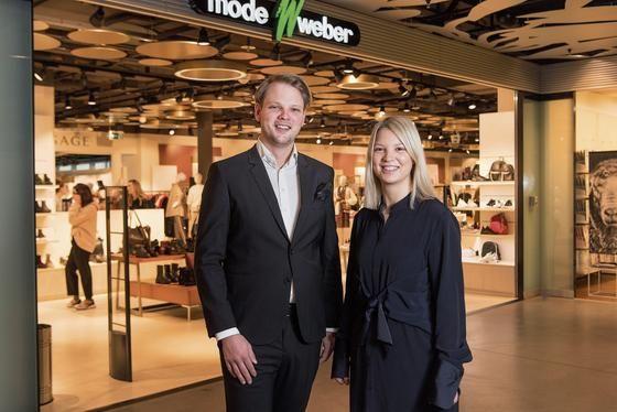A smiling man and woman standing proudly in front of a Mode Weber shoe store entrance.